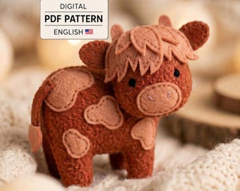 May include: A felt craft toy shaped like a Highland cow with a rust-colored body and light tan patches. The toy features a shaggy fringe, small ears, and black bead eyes. A digital label reads: DIGITAL PDF PATTERN ENGLISH.