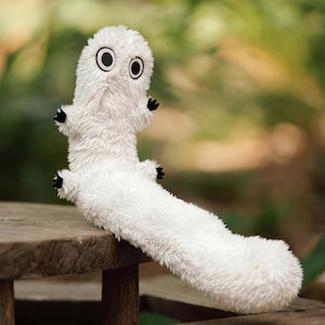 May include: A white plush toy with a long, furry body and a rounded head. It features two large, circular black and white eyes and four small, stubby limbs tipped with black claws. The texture of the fabric is shaggy and soft throughout.