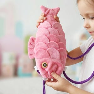 May include: A plush pink fish-shaped bag featuring layered scale-like fabric details, a single round button eye with a dark centre, and a braided purple cord strap. The soft textile construction mimics the form of a fish with a tail and fins.