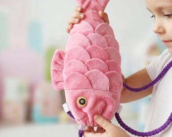 May include: A plush pink fish-shaped bag featuring layered scale-like fabric details, a single round button eye with a dark center, and a braided purple cord strap. The soft textile construction mimics the form of a fish with a tail and fins.