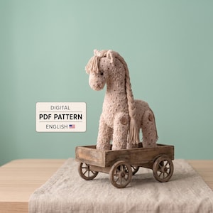 May include: A beige knitted horse toy with a long braided mane and tail, featuring small brown buttons at the joints, standing inside a small wooden four-wheeled cart. A rectangular sign displays the text DIGITAL PDF PATTERN ENGLISH.