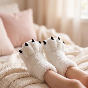Peut inclure: Une paire de chaussons en peluche blanche conçus pour ressembler à des pattes d'animaux. Les chaussons présentent une matière blanche douce et duveteuse avec des sections d'orteils distinctes et des griffes noires pointues sur chaque orteil.
