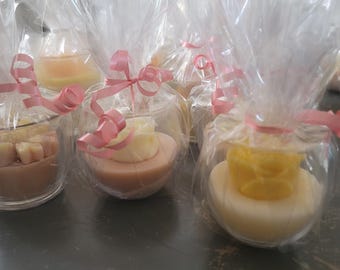 May include: Multiple small, round, clear plastic containers each holding a solid, flat-bottomed wax or soap base topped with a sculpted floral shape in shades of white, yellow, and pink. Each container is wrapped in clear cellophane with a pink ribbon.