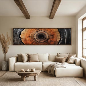 May include: A large, rectangular framed artwork depicting a cross-section of a tree trunk, showcasing concentric rings of orange and brown hues. The artwork hangs above a cream-colored sectional sofa with accent pillows. A wooden coffee table and rug complete the living room setting.