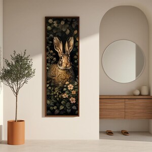 May include: A vertical rectangular wall art print featuring a brown rabbit centered among dark green foliage and small light-colored flowers. The artwork is framed in a thin dark brown border, showcasing a detailed illustration of the animal.