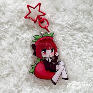 May include: An acrylic keyring featuring a cartoon character with short red hair and a dark dress sitting on a large red strawberry with green leaves. The keyring has a red star-shaped clasp attached to the top of the clear acrylic charm.