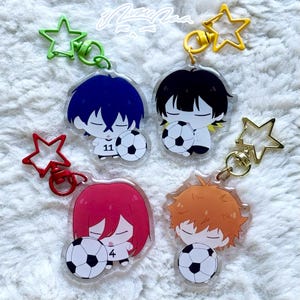 May include: Four anime-style keyrings featuring characters with soccer balls. Each keyring has a different hair colour: blue, black, red, and orange. Each character is holding a soccer ball. The keyrings have star-shaped accents in green, yellow, and red.
