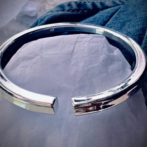 May include: A silver-colored, open-ended cuff bracelet. The bracelet has a smooth, polished surface and a rectangular cross-section. The open ends of the bracelet are squared off. The bracelet is resting on a white surface and blue denim.