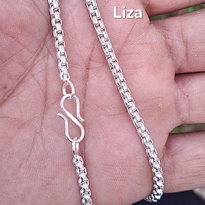 Pode incluir: Um close-up de um colar de prata com um design de corrente de caixa. O colar apresenta um fecho de lagosta e a palavra "Liza" no canto superior direito. A corrente é feita de pequenas ligações quadradas.