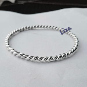 Pode incluir: Uma pulseira prateada, em estilo corda torcida. A pulseira é circular e parece ser feita de um metal brilhante. A palavra "Liza" está escrita em itálico na pulseira. A pulseira está sobre uma superfície branca.