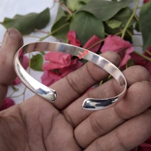 May include: A polished silver cuff bracelet with rounded edges. The open design allows for easy wear. The bracelet is held in a hand, with the word "Liza" visible on the bracelet. The bracelet is a simple, elegant accessory.