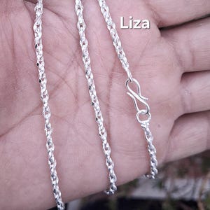 Pode incluir: Um colar prateado com um design torcido e texturizado. O colar tem um fecho de lagosta. A palavra "Liza" está impressa em branco no canto superior direito.