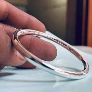 May include: A polished silver bangle bracelet. The round bracelet is smooth and reflective, with a simple, elegant design. The bangle is held by a hand, showcasing its bright, metallic sheen. A classic piece of jewelry.