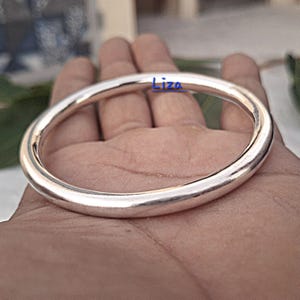 Könnte beinhalten: Ein silberfarbenes Armreif-Armband mit glattem, abgerundetem Design. Das Armband wird in einer Hand gehalten, und das Wort "Liza" ist in Blau auf dem Armband geschrieben. Das Armreif ist ein schlichtes, elegantes Schmuckstück.