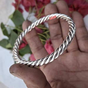 Può includere: Un braccialetto a bracciale argentato con un design a corda intrecciata. Il braccialetto è circolare e sembra essere in metallo. La parola "Liza" è scritta sul braccialetto. Lo sfondo comprende foglie verdi e fiori rosa.