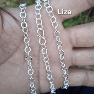Pode incluir: Um colar de corrente de prata com fecho lagosta. A corrente é feita de pequenas elos ovais interligados. A palavra "Liza" está impressa em branco no canto superior direito da imagem.