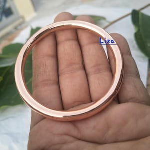 May include: A rose gold bangle bracelet. The bracelet is a solid, circular band with a smooth, polished finish. The color is a warm, metallic rose gold. The bangle is a simple, elegant piece of jewelry.