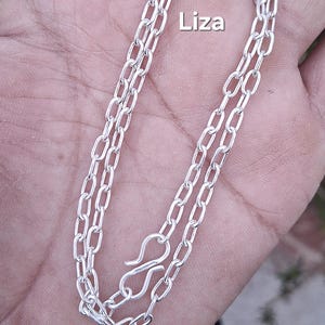 Op de afbeelding: Een zilveren ketting met een delicaat, geschakeld ontwerp. De ketting heeft een haaksluiting en het woord "Liza" in witte tekst. De ketting wordt gepresenteerd tegen een neutrale achtergrond.