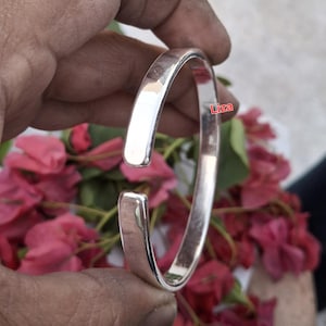 Peut inclure: Un bracelet manchette argenté brillant avec un design lisse et arrondi. Le bracelet ouvert est présenté sur un fond de fleurs roses. Le bracelet est un bijou simple et élégant.
