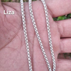Può includere: Una collana a catena di tipo box color argento con un motivo a maglie quadrate ripetute. La parola Liza è stampata in bianco sul lato sinistro dell'immagine. La catena è composta da maglie metalliche lucide che formano un filo uniforme.