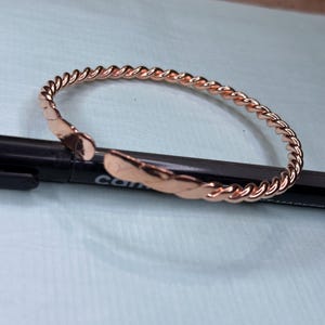 May include: A rose gold-colored, open cuff bracelet with a twisted rope design. The bracelet is made of a shiny metal and has a hammered texture on the ends. The bracelet is approximately 2.5 inches in diameter.