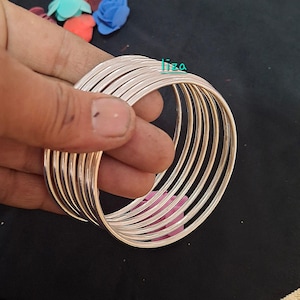 Peut inclure: Plusieurs bracelets de couleur argentée. Les bracelets sont fins et ronds, avec une surface lisse et réfléchissante. Ils sont tenus dans une main, sur un fond sombre. Le mot "liza" est écrit en vert en haut à droite.