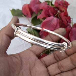 May include: A silver adjustable bangle bracelet. The bracelet has a smooth, polished finish and a simple design. The bracelet is held in a hand, with a background of pink flowers.
