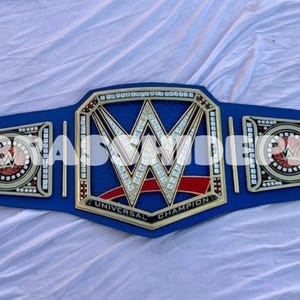May include: A blue and gold championship belt with the letters "W" in the center. The belt has a blue strap and gold accents. The words "UNIVERSAL CHAMPION" are visible.