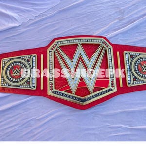 Puede incluir: Un cinturón de campeonato rojo y dorado con el logotipo de la WWE en el centro. El cinturón tiene detalles dorados y las palabras "UNIVERSAL CHAMPION" son visibles. El cinturón está sobre una superficie blanca.