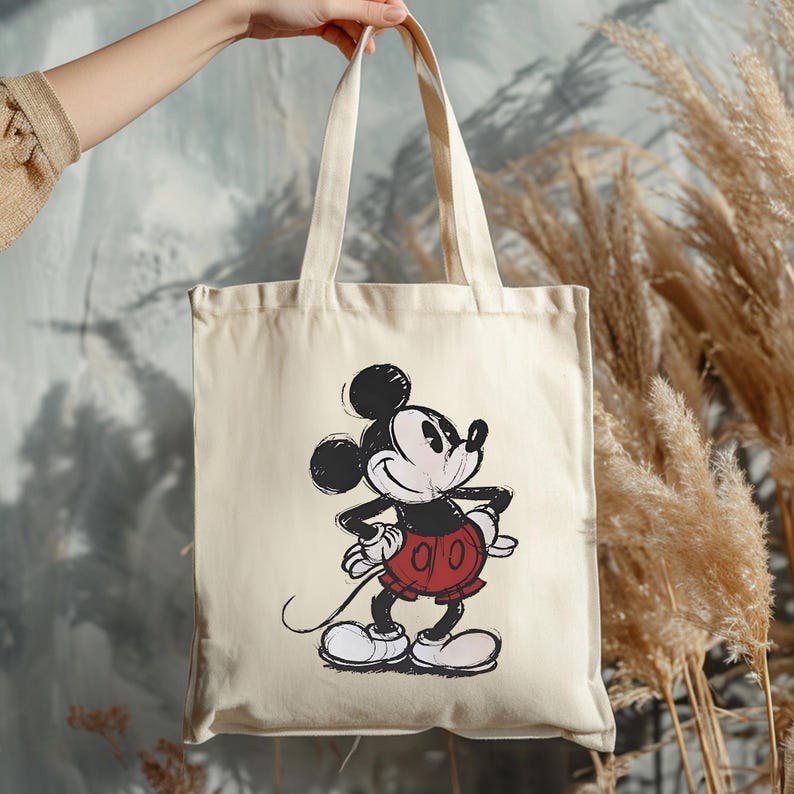 Puede incluir: Bolso tote de color crema con una ilustraci&oacute;n cl&aacute;sica de Mickey Mouse. El personaje de dibujos animados est&aacute; representado en negro, rojo y blanco, con un estilo de boceto. El bolso tiene asas largas y lo sostiene una persona.
