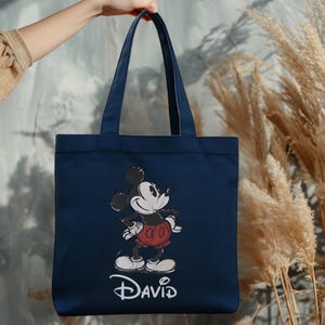 Puede incluir: Bolso tote azul marino con un dise&ntilde;o de Mickey Mouse. El personaje de dibujos animados es de color negro, blanco y rojo, con el nombre "David" en escritura blanca. El bolso tiene dos asas y se sostiene.