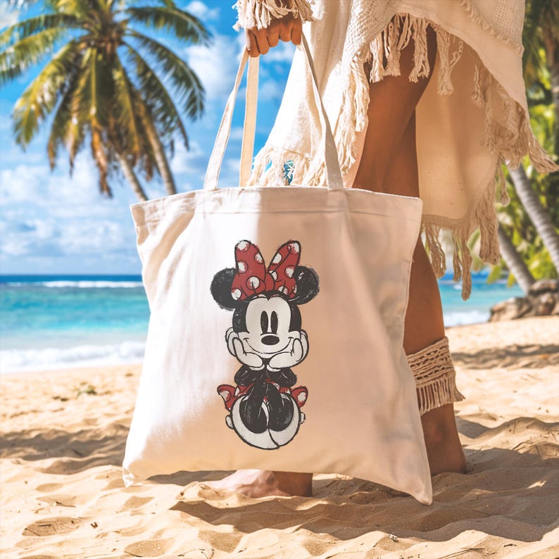 Puede incluir: Un bolso tote de color crema con un dise&ntilde;o de Minnie Mouse. El personaje de dibujos animados est&aacute; en negro, rojo y blanco, con un lazo de lunares. El bolso tiene asas largas de color crema y se sostiene en una playa de arena con palmeras y el oc&eacute;ano de fondo.