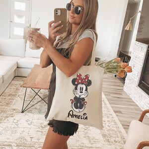 Puede incluir: Bolso tote de color crema con un dise&ntilde;o de Minnie Mouse, que incluye un lazo rojo de lunares y detalles a juego. El nombre "Amelia" est&aacute; impreso debajo. La bolsa est&aacute; llena de tulipanes naranjas.