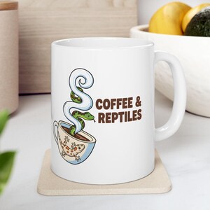 May include: White ceramic mug with a whimsical design. The mug features a cartoon coffee cup with a gecko on the side and a snake emerging as steam. The text "COFFEE & REPTILES" is printed in brown.