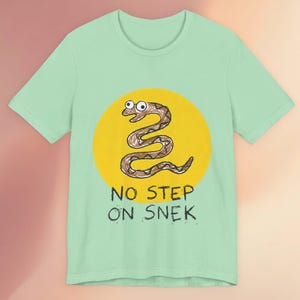 Puede incluir: Camiseta verde claro con un diseño de serpiente de dibujos animados dentro de un círculo amarillo. La serpiente marrón está dibujada con un estilo caprichoso. Debajo de la serpiente, el texto dice "NO STEP ON SNEK" en negro.