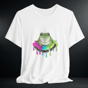 May include: White t-shirt featuring a cartoon frog with the text "Serotonin Dump" in a colorful, dripping font. The frog is green and sits on a multi-colored base of pink, yellow, blue, and green.