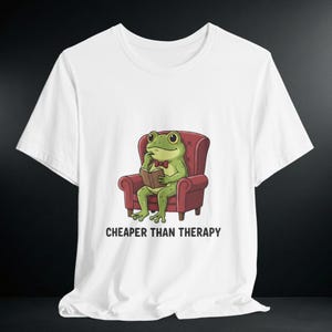 Könnte beinhalten: Weißes T-Shirt mit einem Cartoon-Frosch in einem roten Sessel, der ein Buch liest und eine Fliege trägt. Der Text "CHEAPER THAN THERAPY" ist darunter gedruckt. Das T-Shirt ist aus weichem Material.