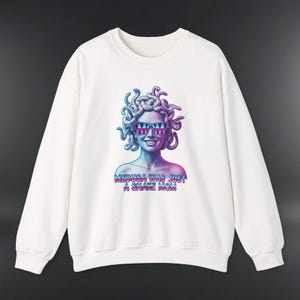 Puede incluir: Sudadera blanca con un gráfico de Medusa con gafas de sol, en tonos azules, morados y rosas. El texto "MEDUSA WAS JUST A SNAKE MOM" está debajo. La sudadera tiene cuello redondo y mangas largas.