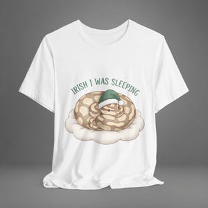 May include: White t-shirt with a cartoon snake wearing a green sleeping cap, curled up on a cloud. The text "Irish I was sleeping" is arched above the snake. The design is centered on the shirt.
