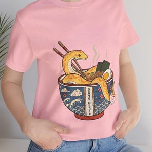 Op de afbeelding: Roze t-shirt met een cartoon illustratie van een gele slang in een ramenkom. De kom heeft een blauw-wit ontwerp met de tekst "Danger Noodle". Eetstokjes, zeewier en een ei zitten ook in de kom.