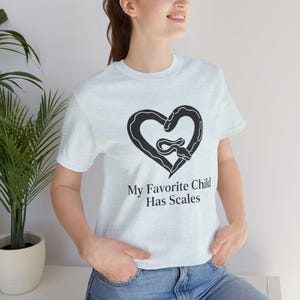Op de afbeelding: Lichtblauw T-shirt met een zwart hartvormig ontwerp met een slang. De tekst "My Favorite Child Has Scales" staat eronder. Een casual kledingstuk.