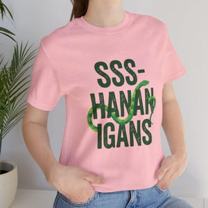 May include: A pale pink t-shirt featuring the words "SSS-HANAN-IGANS" in dark green, with a green snake coiled around the word "HANAN". The shirt is made of a soft material.