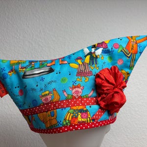 Könnte beinhalten: Ein skurriler, blauer Stoffhut mit einem verspielten Design mit Cartoon-Schweinen, Noten und Rollschuhen. Ein rotes Band und eine große, rote Stoffblume schmücken den Hut. Der Hut hat ein rotes Etikett mit MyMutzje.de.