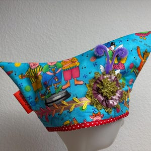 Könnte beinhalten: Ein skurriler, spitzer Hut mit einem leuchtend blauen Hintergrund, der Cartoon-Charaktere beim Musizieren zeigt. Der Hut ist mit lila Pompons, einer gold-violetten Blume und einer roten Quaste verziert. Der Rand ist mit roten Punkten versehen.