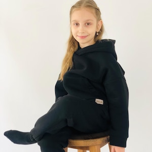 May include: A person wearing a black hoodie and matching tracksuit bottoms, seated on a wooden stool. The outfit features a small label with text. The background is a plain white wall.
