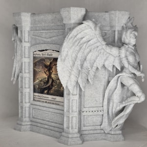 May include: A light gray speckled stone-textured card display stand featuring architectural pillar details and a sculpted winged figure on the side. A Magic: The Gathering trading card titled Iona, Shield of Emeria is inserted into the front slot.