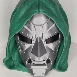 Op de afbeelding: Een zilverkleurig masker met een metallic afwerking, voorzien van een stijf, geklonken ontwerp met zwarte oog- en mondopeningen. Het masker wordt omlijst door een getextureerde, donkergroene kap die over de bovenkant en zijkanten valt.