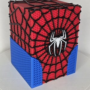 Puede incluir: Una caja de almacenamiento cuadrada con una superficie de panal roja y azul. La parte superior y los lados presentan un patrón de telaraña negra en relieve, y un emblema de araña blanco está centrado en el panel frontal rojo.