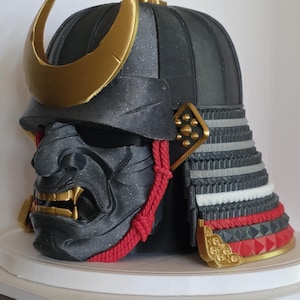 Könnte beinhalten: Schwarzer und goldener Samurai-Helm mit furchterregender Gesichtsmaske. Der Helm hat einen goldenen Halbmond, rote Seilakzente und goldene Details. Der Nackenschutz ist geschichtet mit roten und weißen Abschnitten und zeigt detaillierte Handwerkskunst.