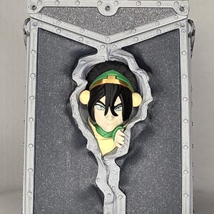 May include: A gray rectangular box featuring a 3D sculpted image of a character with black hair, a green headband, and a yellow and green outfit. The character is emerging from a torn section of the box.